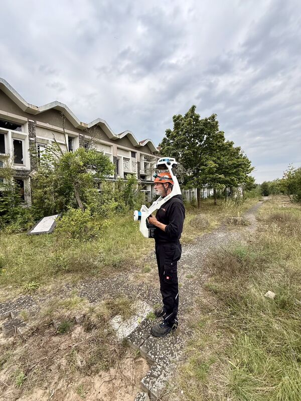 bgis-Mitarbeiter mit Schutzhelm erfasst mit dem Mobile-Mapping-System NavVis VLX die Fassade und das Gelände eines leerstehenden Bestandsgebäudes