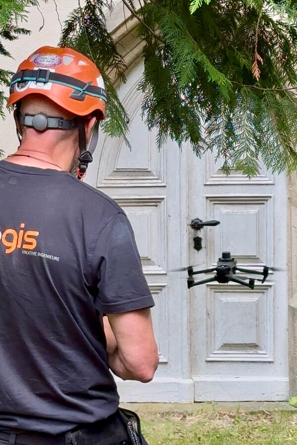 bgis-Mitarbeiter mit Schutzhelm steuert eine DJI-Drohne vor dem historischen Holzportal eines denkmalgeschützten Gebäudes